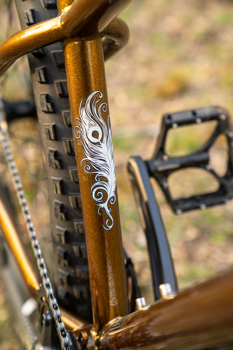 Close-up of a bicycle frame featuring a shimmering metallic bronze finish, adorned with an intricate white feather design. The bike's rear tire and part of the pedal are visible, set against a blurred natural background.