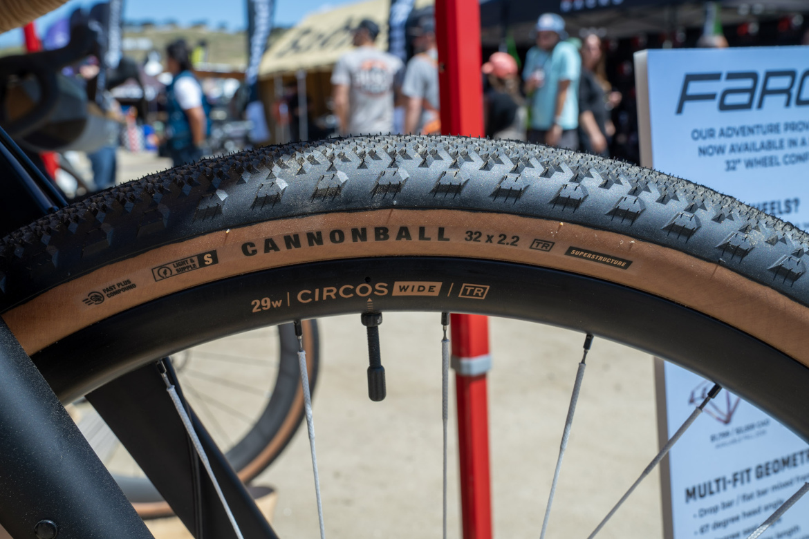 Close-up of a bicycle tire featuring the inscription "CANNONBALL 32 x 2.2" and "29w CIRCOS WIDE TR" on the sidewall. The tire has a textured tread pattern and a tan sidewall, set against a blurred outdoor background with people and vendor tents at a cycling event.
