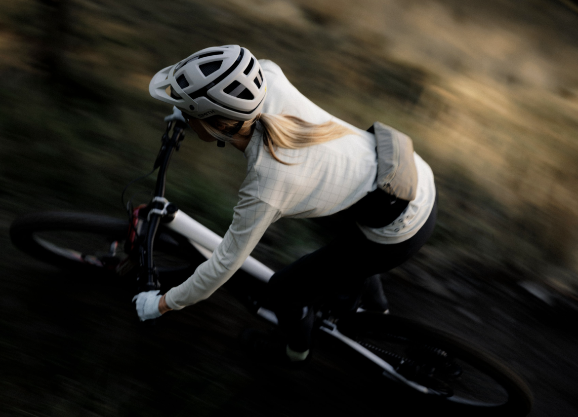 A person riding a mountain bike on a trail, captured in motion. The cyclist is wearing a helmet and a lightweight, long-sleeve jersey, with their blonde hair flowing behind them. The background is blurred, suggesting speed and movement through a wooded area.