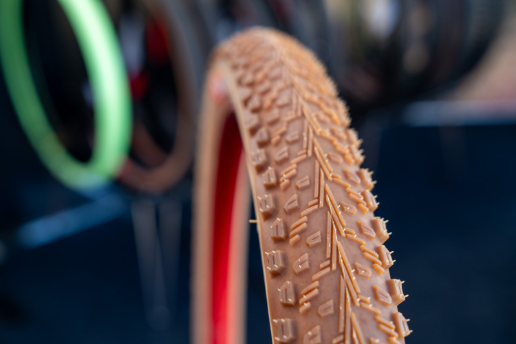 Close-up view of a bicycle tire with a textured tread pattern, showcasing its grooves and ridges. The tire is brown with a red lining, and several other colorful tires are slightly blurred in the background.