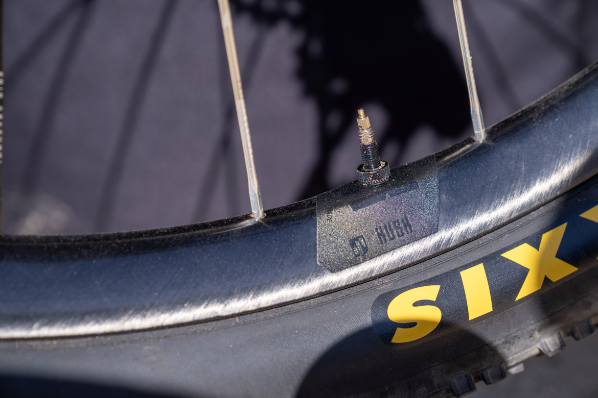 Close-up of a bicycle wheel rim featuring a valve stem and a label displaying "HUSH," with a black rim and yellow "SIX" branding visible on the tire. The background is softly blurred, emphasizing the details of the wheel.