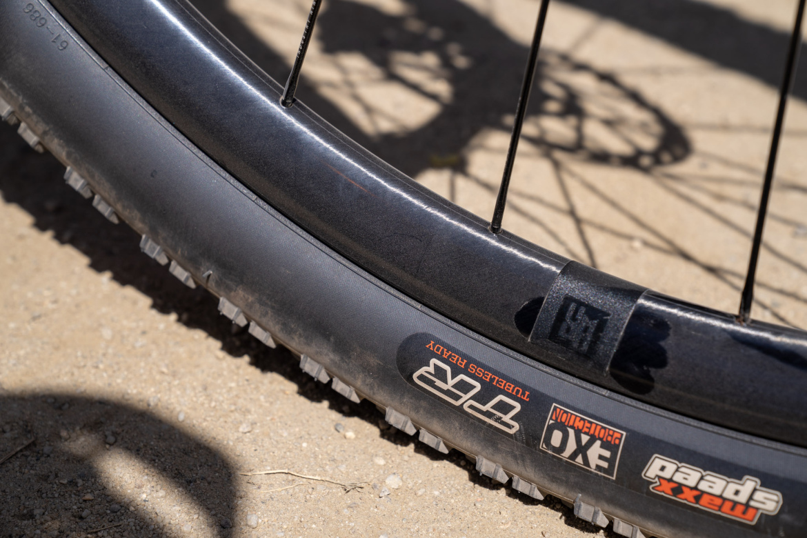 Close-up view of a mountain bike tire and wheel, showing the textured surface of the tire tread and the wheel's rim details, resting on a sandy surface. The tire has visible branding and labels indicating it's tubeless ready.