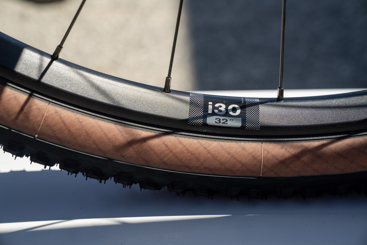 Close-up of a bike wheel rim showing a black surface with a silver label marked "i30" and "32". The rim features a wood-like finish on the inner side, with visible spokes extending towards the hub. The background is blurred, emphasizing the details of the wheel.