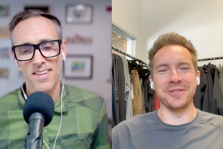 Two men are engaged in a video conversation. The man on the left, wearing glasses and a green plaid shirt, speaks into a microphone. The man on the right, smiling and wearing earbuds, is dressed in a gray shirt and is in a space with clothing hanging on racks. The setting appears casual and friendly.