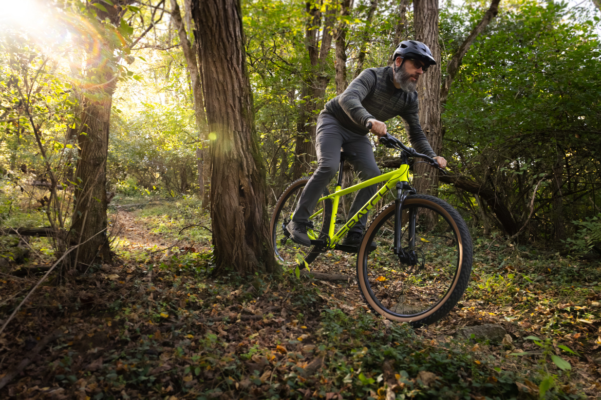 A man with a beard wearing a helmet and outdoor gear rides a bright yellow mountain bike through a wooded area, surrounded by trees and dappled sunlight filtering through the leaves.