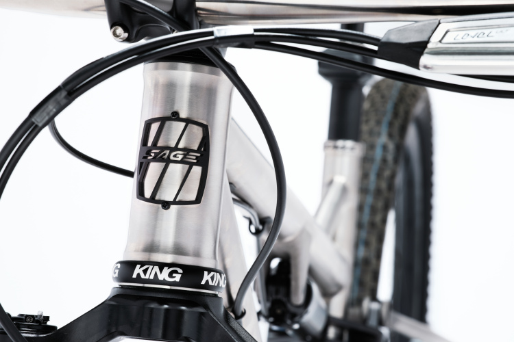 Close-up image of a bicycle's head tube featuring a stainless steel frame with a "SAGE" logo. The image also shows the handlebars, integrated cables, and a "KING" logo on the bottom bracket. The background is plain white, emphasizing the bike's components.