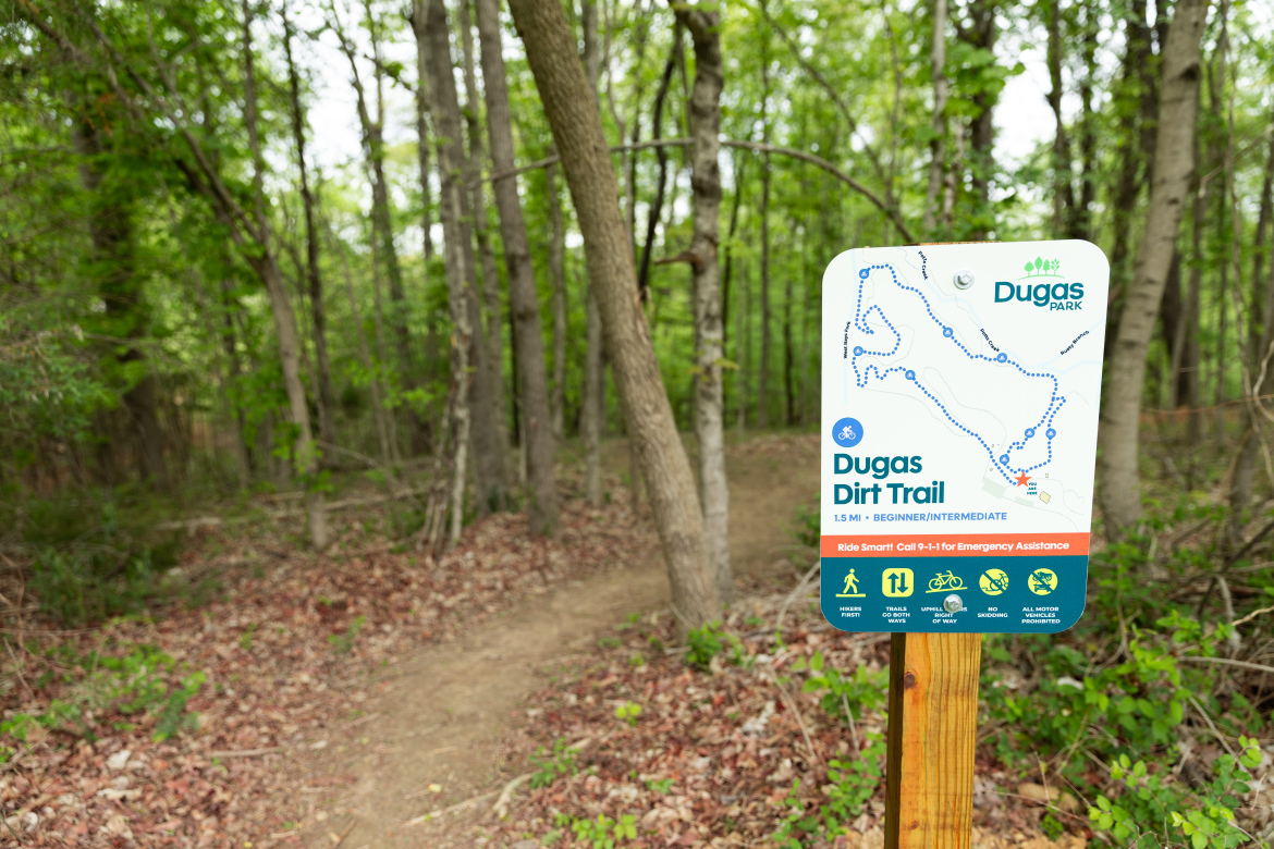 A sign for the Dugas Dirt Trail, indicating a 1.5-mile route suitable for beginner to intermediate riders. The sign features a map of the trail, along with safety instructions and contact information for emergency assistance. The trail is surrounded by lush green trees and natural foliage, with a dirt path visible in the background.