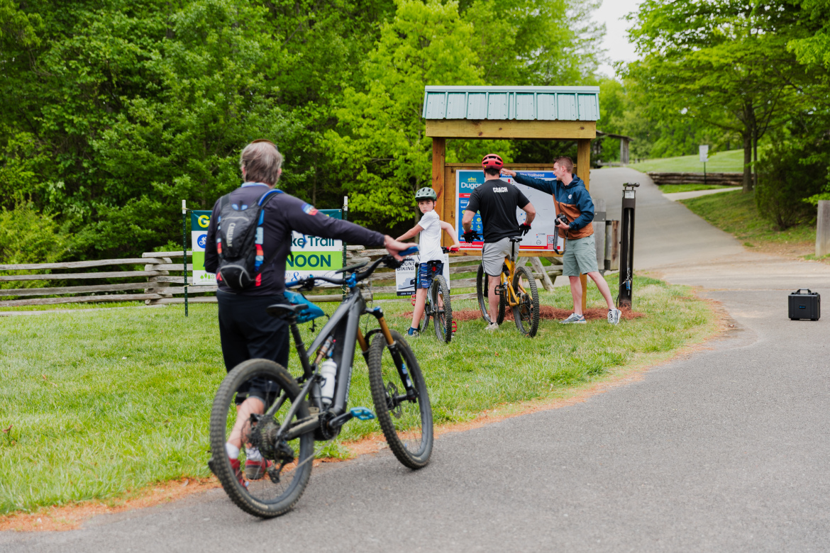 A group of people at a bike trail kiosk, with a sign indicating a guided bike trail starting at noon. One person is wearing a helmet and standing next to a bicycle, while another is speaking to a child and an adult. The scene is surrounded by greenery, with a path leading away from the kiosk.