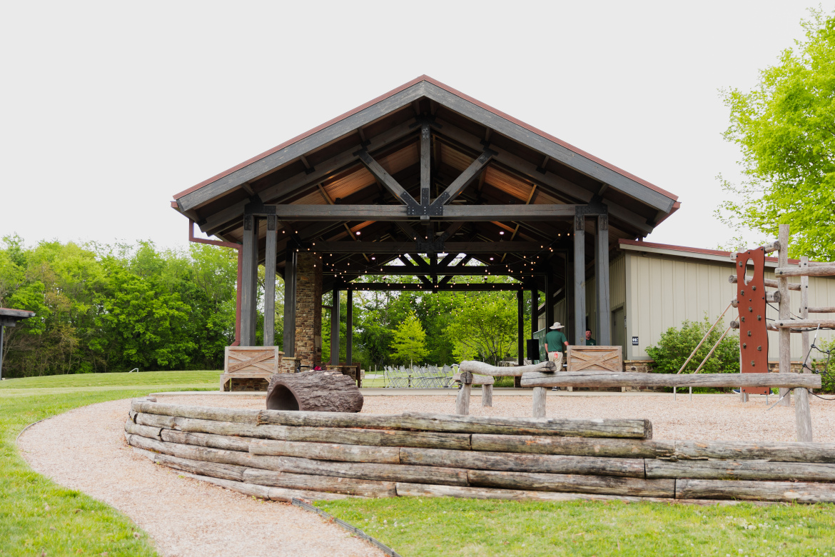 A rustic outdoor pavilion with a peaked roof and wooden beams, illuminated by string lights, surrounded by green trees. In the foreground, a winding gravel path leads to a wooden seating area and playground equipment, while two people are visible near the pavilion.