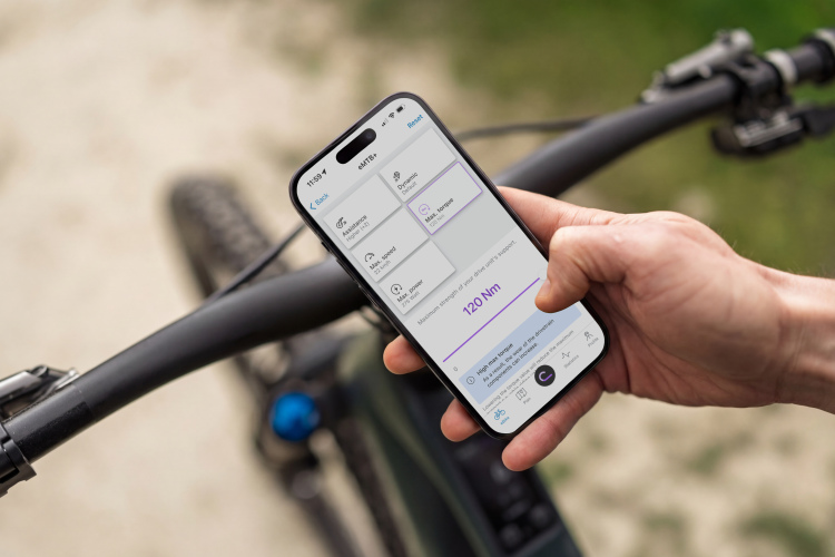 A person holds a smartphone displaying an app interface related to electronic mountain biking, showing settings for assistance levels and maximum torque. The phone is positioned above the handlebars of a bicycle, with a blurred outdoor background.