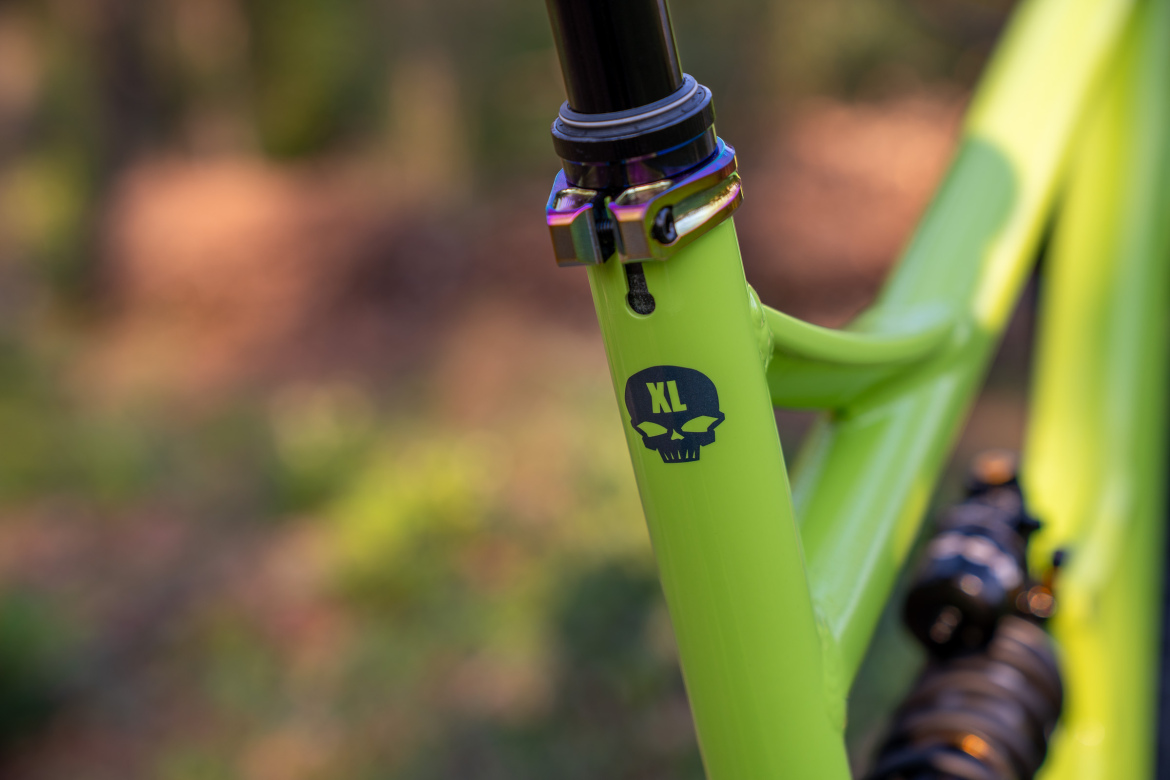 Close-up of a bright green bike frame featuring a black skull logo with "XL" text, showcasing a seat post clamp and a section of the rear suspension system, set against a blurred natural background.