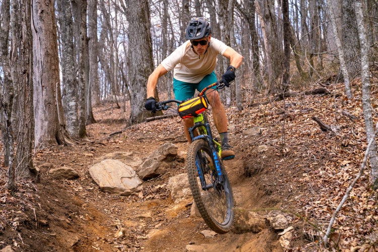 A mountain biker with a black helmet and sunglasses rides down a rocky, wooded trail. The cyclist wears a light-colored shirt and turquoise shorts, leaning forward as they navigate the terrain. Surrounding trees are bare, and the ground is covered in leaves and rocks.