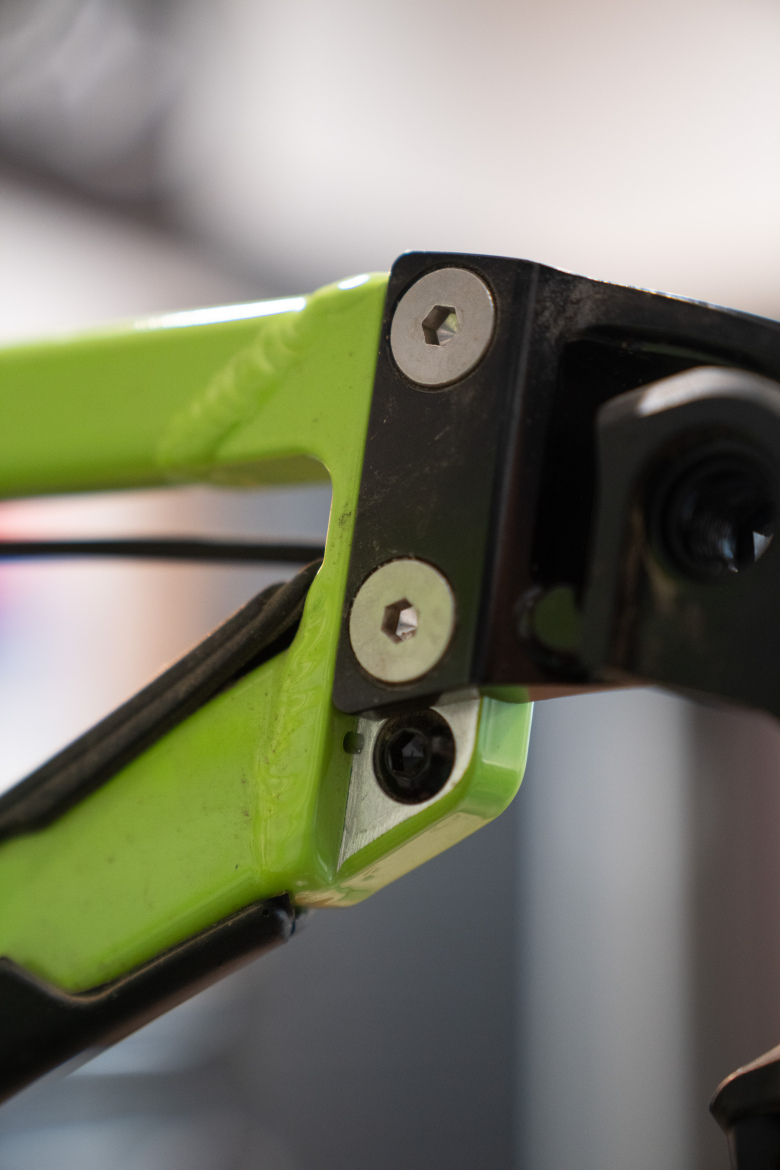 Close-up of a bicycle frame joint featuring a bright green section and metal components, including screws and bolts, indicative of the mechanical structure. The background is blurred, drawing focus to the intricate details of the frame connection.