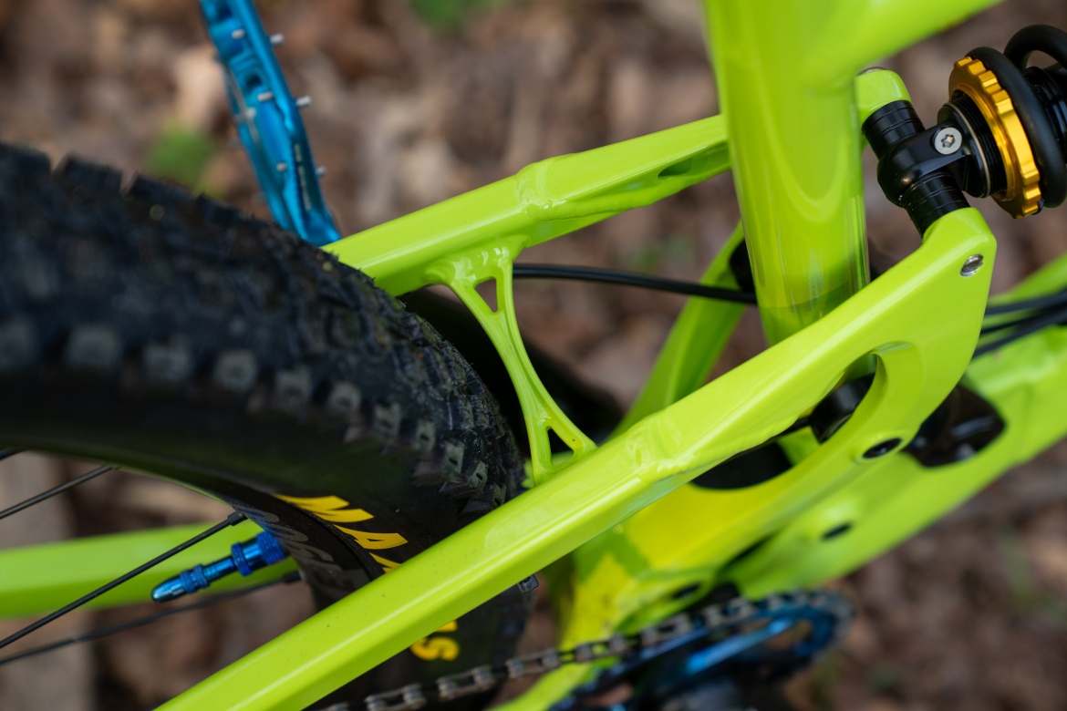 Close-up view of a vibrant green bicycle frame, showcasing detailed suspension components and the textured surface of the tire. The background features soft, blurred natural elements, emphasizing the bike's design.