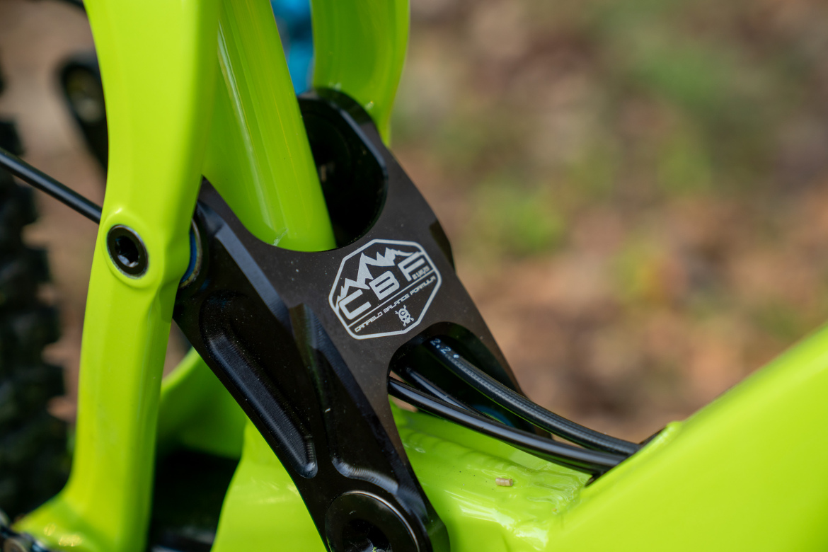 Close-up of a bright green bicycle frame with a black suspension linkage. The linkage features a logo labeled "CBR" and is connected to cables running along the frame. The background is blurred, showing a natural setting.
