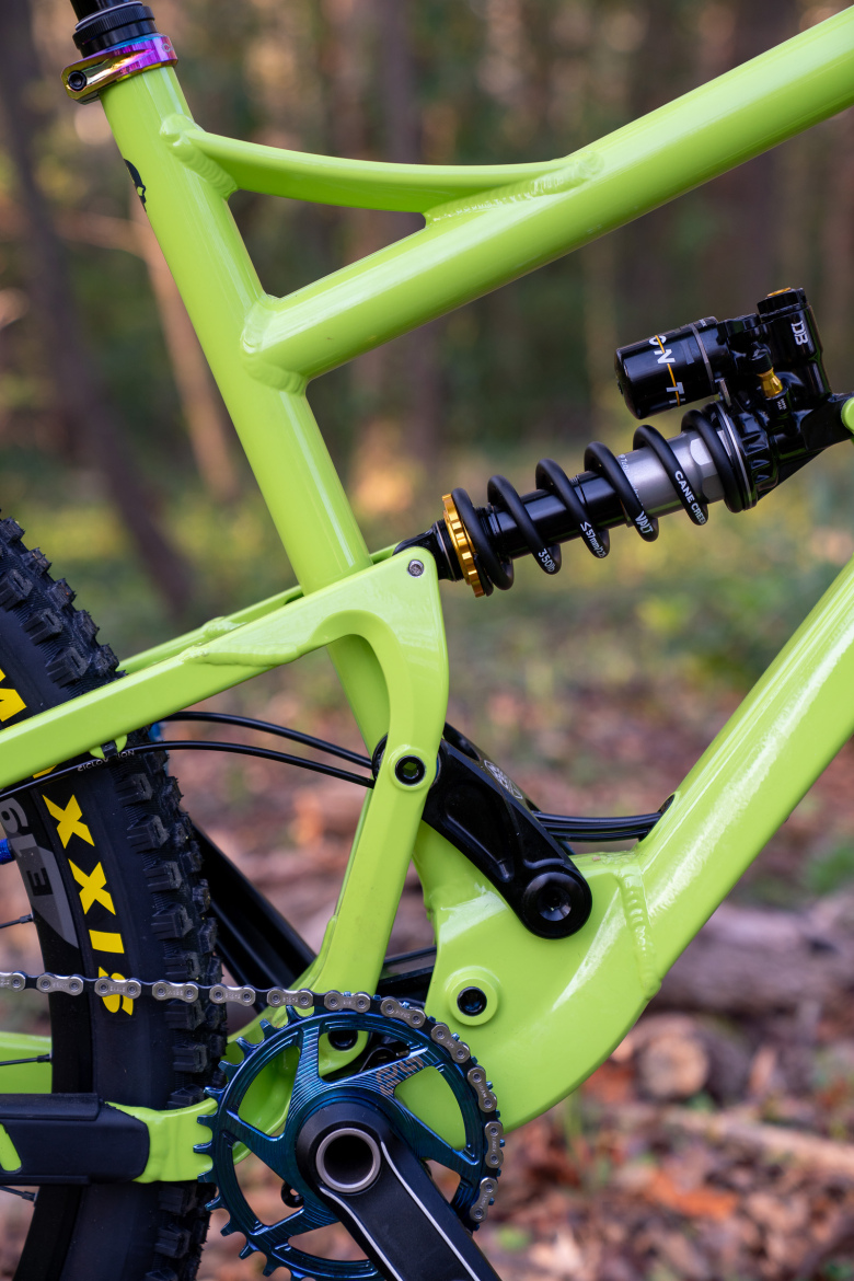Close-up view of a vibrant lime green mountain bike frame featuring a rear shock and suspension system. The image highlights intricate details of the bike's design, including a blue chainring and black tire tread, set against a blurred natural background with trees and foliage.