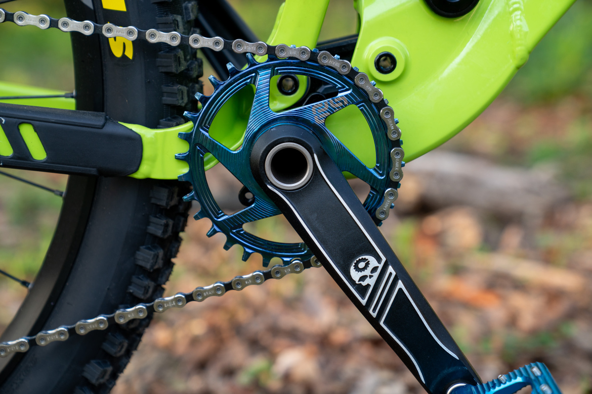Close-up of a mountain bike's crankset and chain, featuring a blue gear with a textured design and a black crank arm adorned with a stylized skull logo. The bike's bright green frame is visible in the background, set against a natural, leaf-covered ground.
