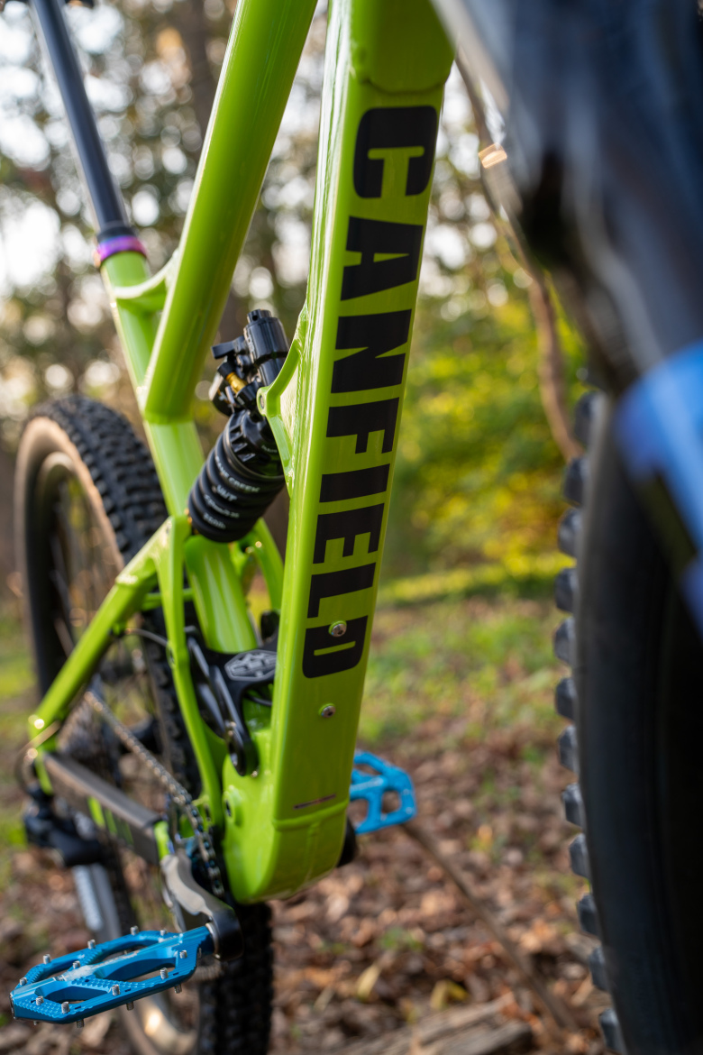An angled close-up of a vibrant green mountain bike frame featuring the name "CANFIELD" prominently displayed. The image highlights the bike's rear suspension and blue pedals against a blurred natural background with trees and leaves.