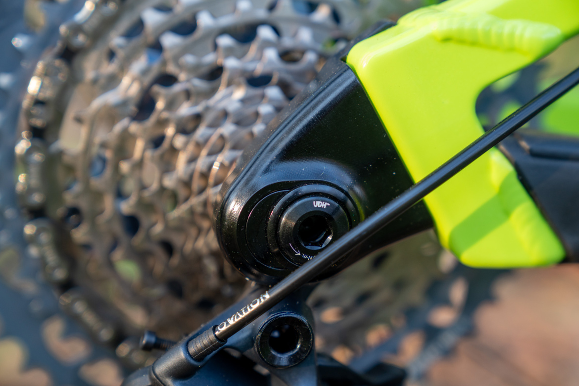 Close-up image of a bicycle's rear derailleur, featuring a black and green component with a visible adjustment dial labeled "UDH," alongside a silver chain and cassette in the background.