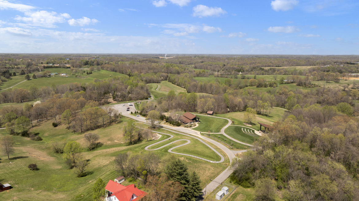 Aerial view of a sprawling green landscape featuring rolling hills, wooded areas, and a winding road. In the foreground, there is a parking area with several cars, and a building with a red roof. The scene is dotted with trees in various stages of foliage, under a bright blue sky with scattered clouds.