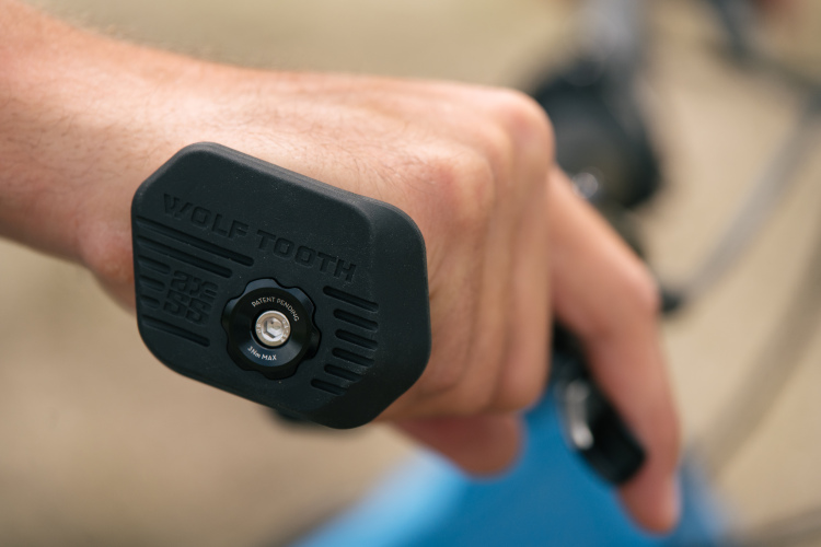 A close-up of a hand gripping a black Wolf Tooth component attached to a bicycle handlebar, featuring a circular adjustment dial marked with "PATENT PENDING" and a torque limit indicator. The background shows parts of a bicycle, suggesting an outdoor setting.