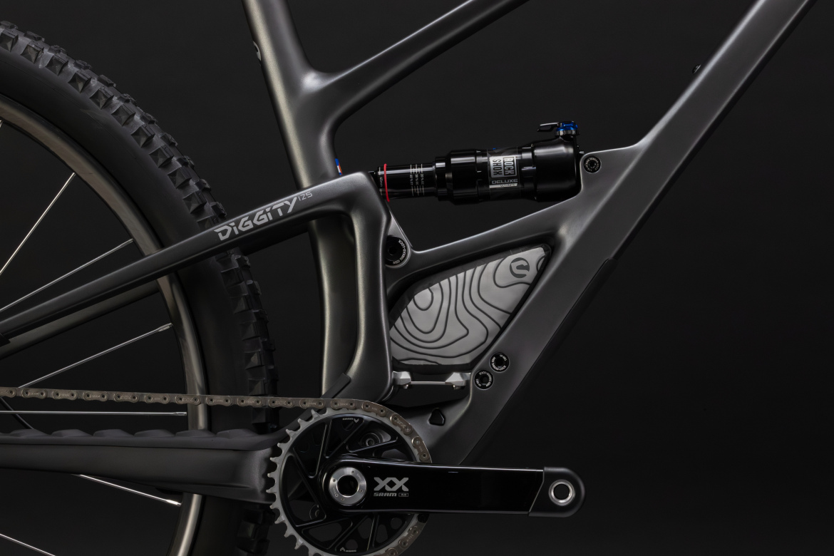Detailed close-up of a modern mountain bike frame, showcasing the suspension system and drivetrain components, against a black background. The frame features sleek lines and a subtle gray finish, with a visible shock absorber and gear system.