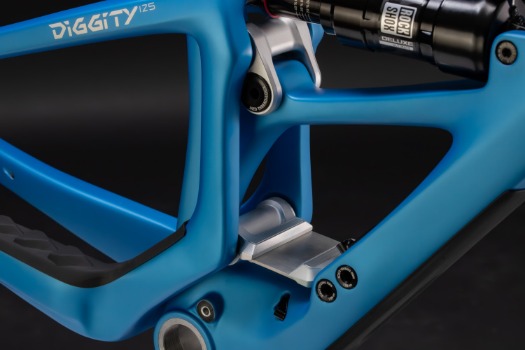 A close-up view of a blue bicycle frame featuring the name "DIGGITY" and details of its shock mount and linkage system. The image highlights the smooth curves and modern design elements, showcasing a combination of aluminum and composite materials with a sleek matte finish, set against a dark background.