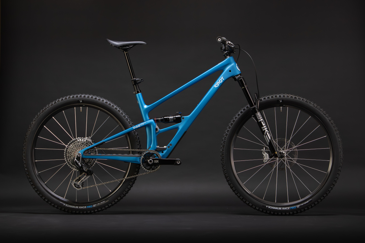 A blue mountain bike positioned against a dark background, featuring a lightweight frame, thick tires for off-road traction, and high-quality components including a rear shock and a visible drivetrain. The bike is angled to display its design and features prominently.