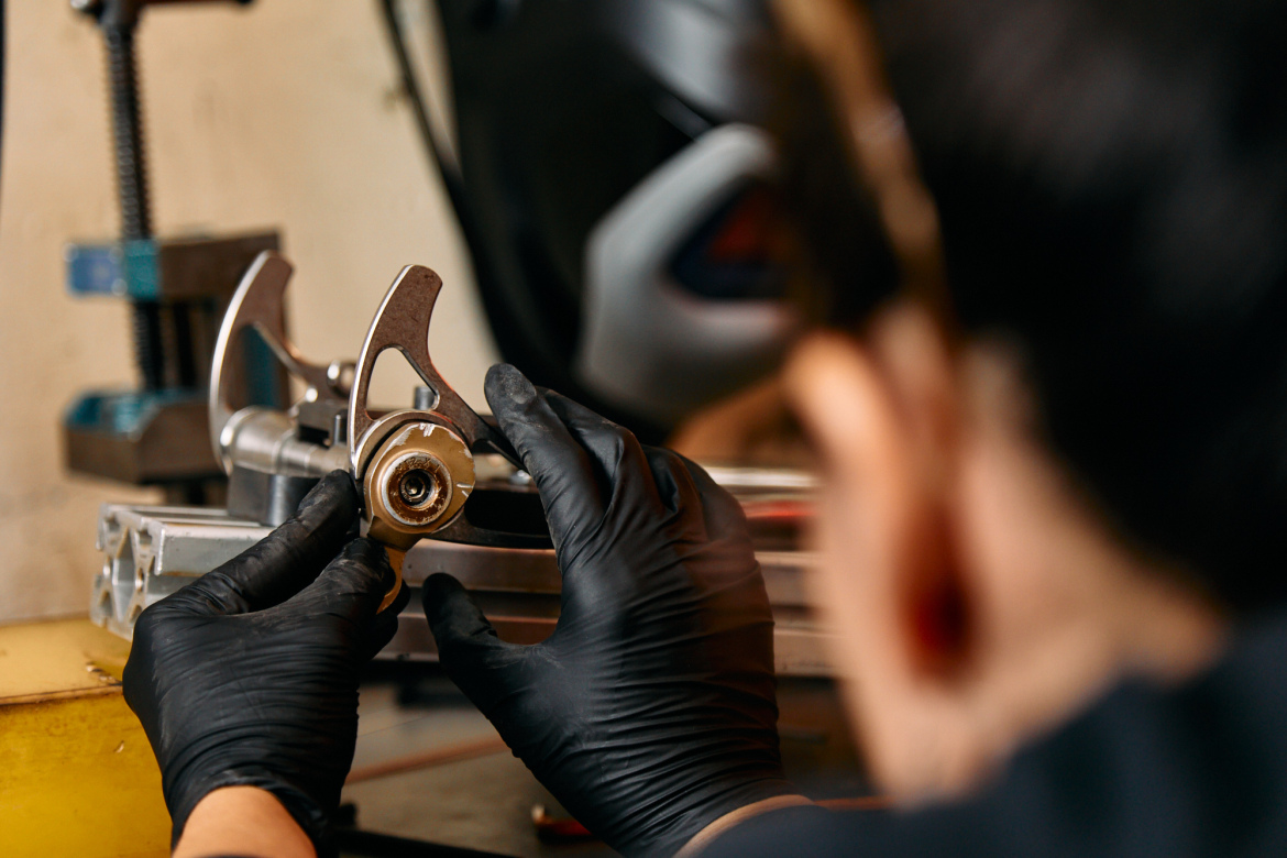 A person wearing black gloves is closely examining and handling a mechanical component, possibly part of a machine or vehicle, while working on a workshop table with various tools nearby.