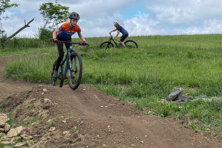 Two young riders are mountain biking on a dirt trail surrounded by grassy hills. The rider in the foreground, wearing a bright orange and blue jersey and a helmet, is jumping off a small dirt mound. In the background, another rider, clad in a dark shirt and helmet, is cycling up the hill. The sky is partly cloudy, creating a vibrant outdoor scene.