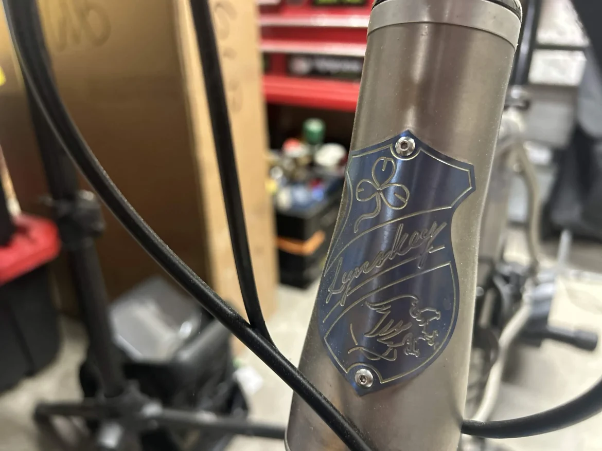 Alt text: Close-up of a bicycle head tube featuring a metal badge with the name "Lynskey" and decorative designs, including a four-leaf clover and a mythical creature. Background shows workshop tools and storage.