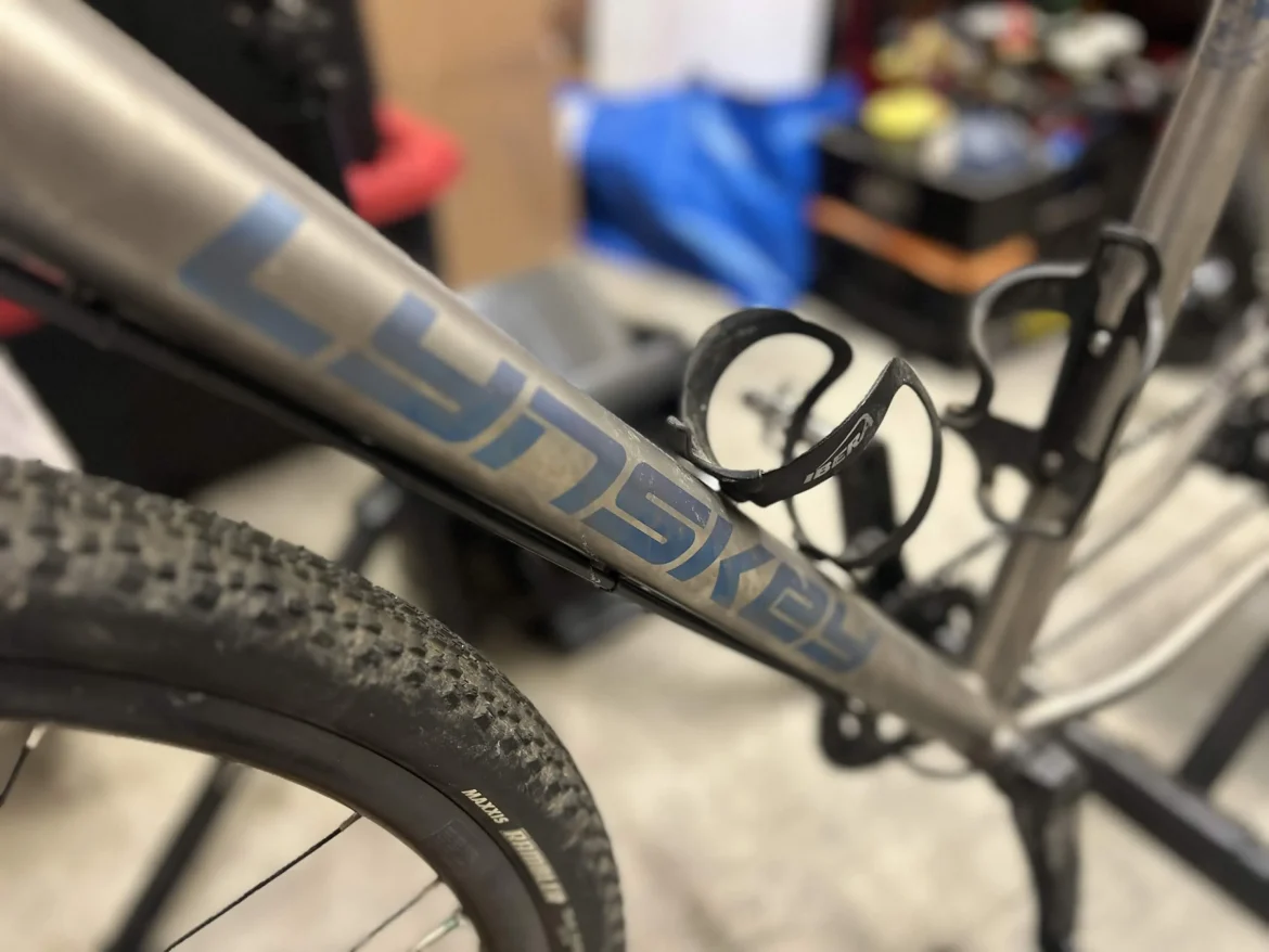 A close-up image of a Lynskey bicycle frame, showcasing the brand logo in blue against a metallic silver surface. The frame features two water bottle holders. In the background, there are blurred objects typical of a workshop setting.