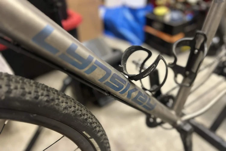 Alt text: Close-up of a bicycle head tube featuring a metal badge with the name "Lynskey" and decorative designs, including a four-leaf clover and a mythical creature. Background shows workshop tools and storage.
