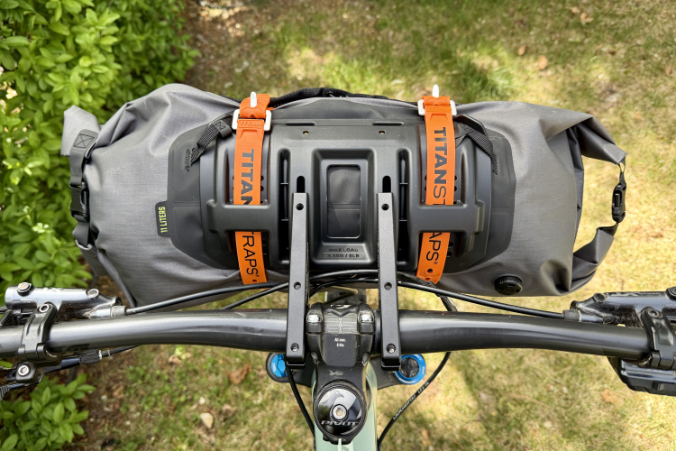 Close-up view of a bicycle handlebar equipped with a mounted bag, secured with orange straps. The bag is gray and designed for outdoor use. The background features blurred greenery, indicating a natural setting.