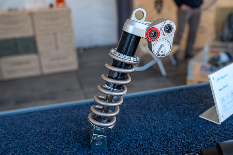A close-up of a mountain bike rear shock absorber displayed on a blue fabric surface, featuring a metal coil spring and adjustable damping controls. In the background, there are blurred boxes and other equipment, indicating a showcase or exhibition setting.