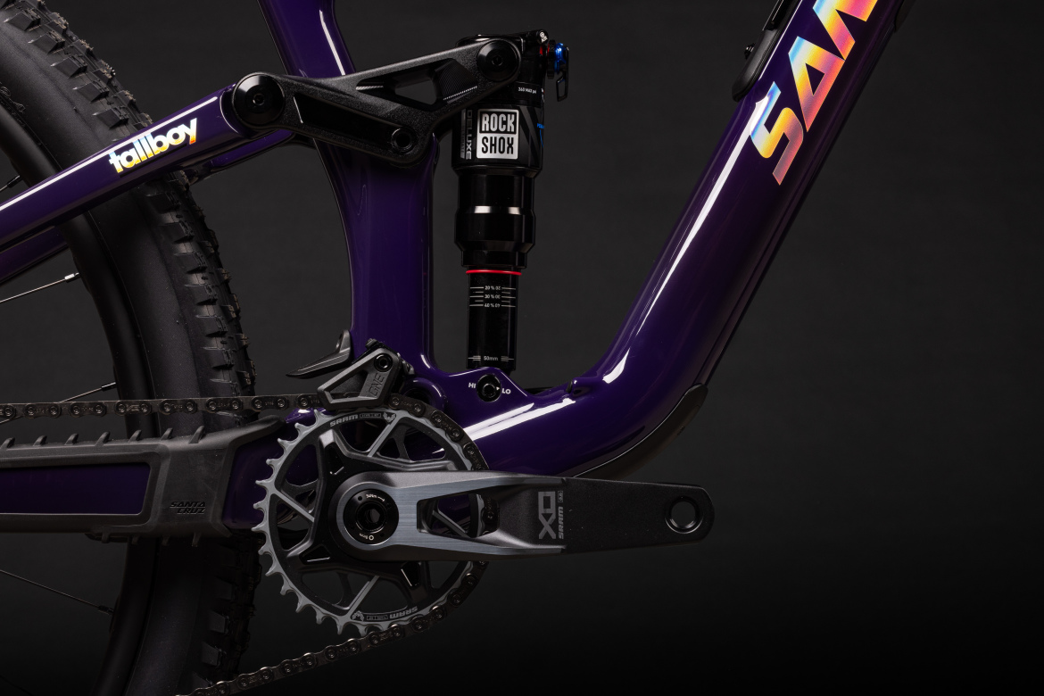 Close-up view of a purple mountain bike frame showcasing the crankset, chainring, and rear shock absorber. The bike features a sleek design with components including a Rock Shox shock and SRAM drivetrain. The background is black, emphasizing the bike's vibrant colors and details.