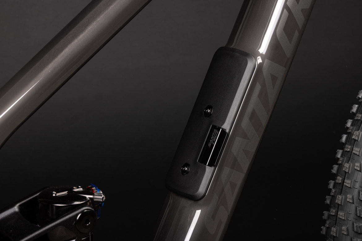Close-up view of a mountain bike frame showcasing the sleek gray finish, featuring a black component with branding, positioned on the underside of the top tube near the rear suspension area. Nearby, a portion of the textured bicycle tire is visible. The background is a solid dark color, enhancing the bike's design details.