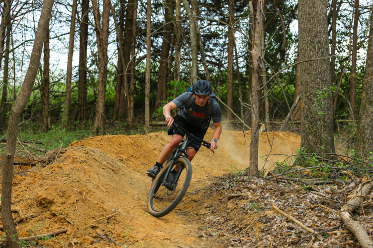 A mountain biker navigates a dirt trail in a wooded area, leaning into a turn as dust kicks up from the ground. The surrounding trees provide a natural backdrop, indicating an outdoor adventure in a serene environment.