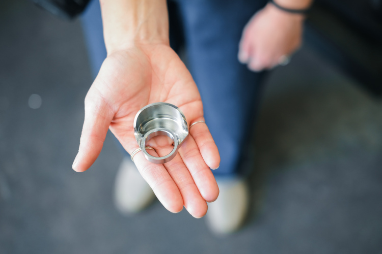 A close-up image of a person's hand holding a shiny, metal ring-shaped object with a hollow center. The person is wearing several rings on their fingers, and the background appears to be a blurred indoor setting.