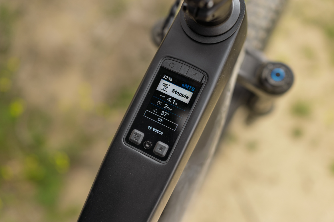 Close-up view of an electric mountain bike's control panel displaying battery level (32%), riding mode (eMTB), and other metrics such as distance (4.1 meters), speed (2 seconds), and incline (37 degrees). The interface features buttons for navigation and a brand logo at the bottom. The background is out of focus, suggesting an outdoor setting.