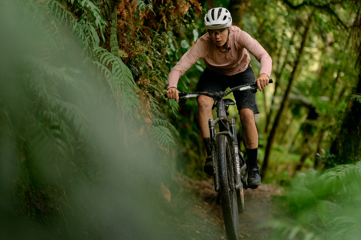 A mountain biker navigating a narrow trail through a lush green forest. The rider is focused and wearing a helmet, with dirt splattered on their clothing from the rugged terrain. Ferns frame the scene, emphasizing the natural setting.