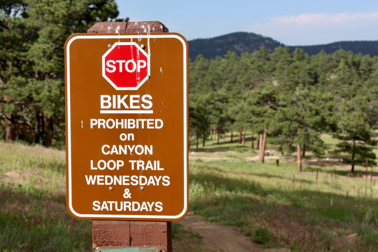 Brown sign with a red stop symbol, indicating that bikes are prohibited on Canyon Loop Trail on Wednesdays and Saturdays, set against a backdrop of green trees and rolling hills.