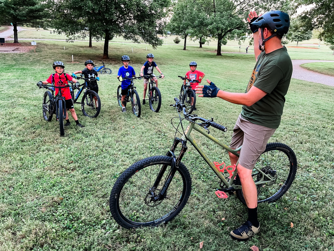 A group of six children in helmets and cycling gear are gathered on a grassy field, each with a mountain bike. An adult instructor, wearing a green shirt and gloves, is standing next to a bike, giving instructions. The background features trees and a grassy path, indicating an outdoor biking environment.