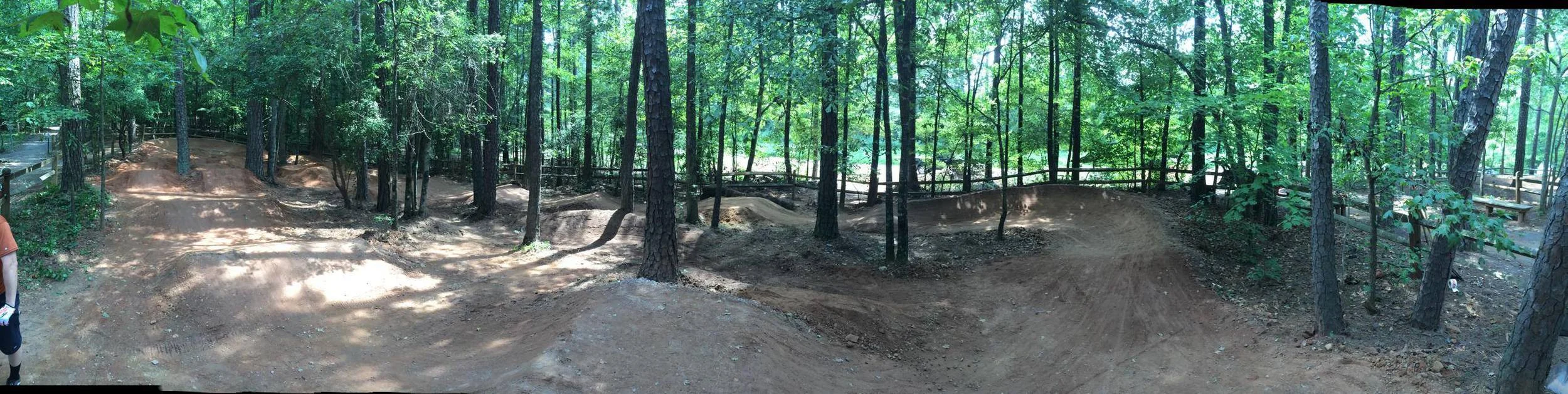 A panoramic view of a dirt bike trail winding through a forested area, featuring various curves and jumps surrounded by greenery and tall trees. The trail is well-maintained, with a natural earthen surface. Wooden fencing is visible in the background, enhancing the outdoor recreational setting.