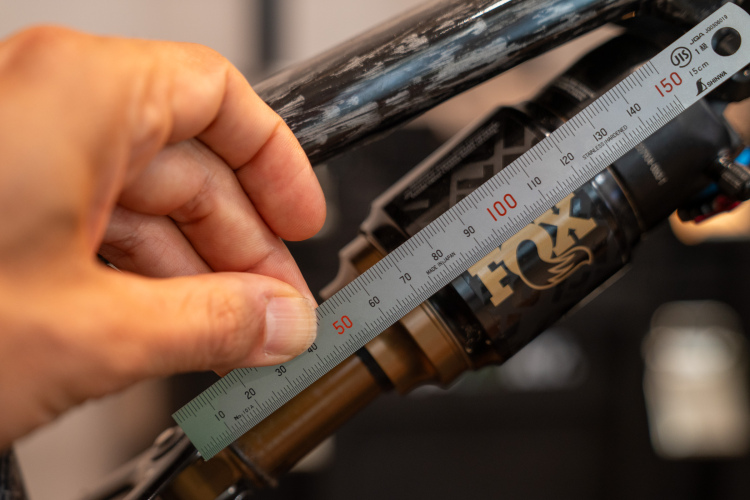 A close-up view of a hand using a ruler to measure a shock absorber component, featuring branding elements. The background is softly blurred, highlighting the measurement process.