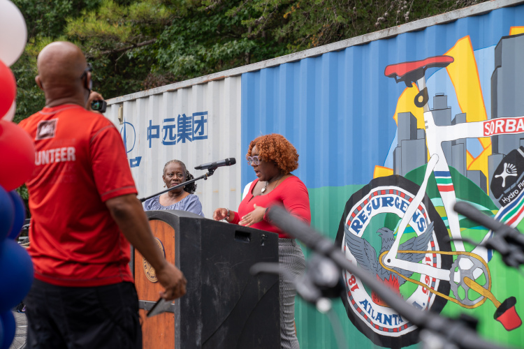 A woman with curly hair in a red top speaks at a podium during an outdoor event, while another woman observes. The background features a colorful mural of a bicycle and city skyline. Balloons in red, white, and blue are visible in the foreground.