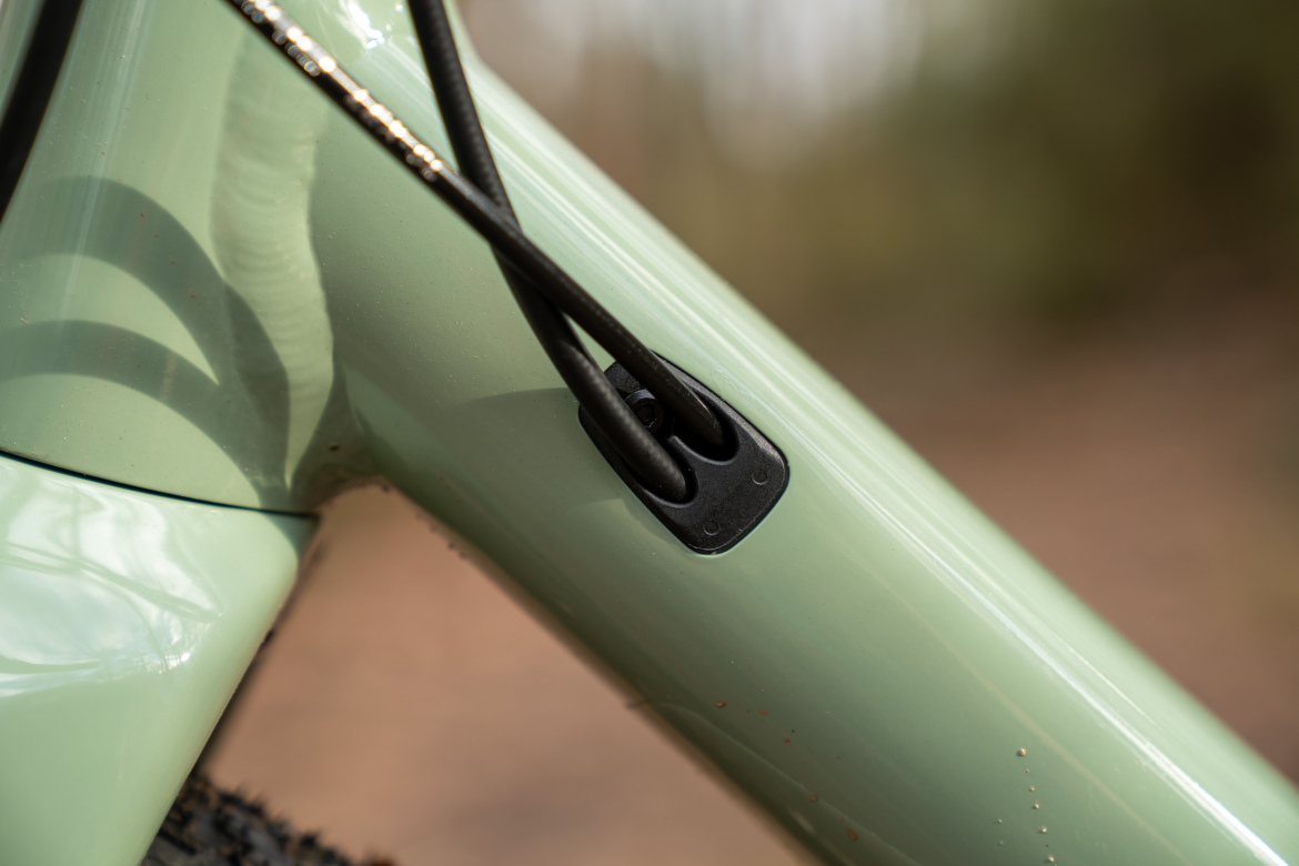Close-up of a bicycle frame showing a green finish and cable routing. The image highlights the connection point where cables are secured, featuring a black rubber grommet. Background is out of focus, suggesting an outdoor setting.