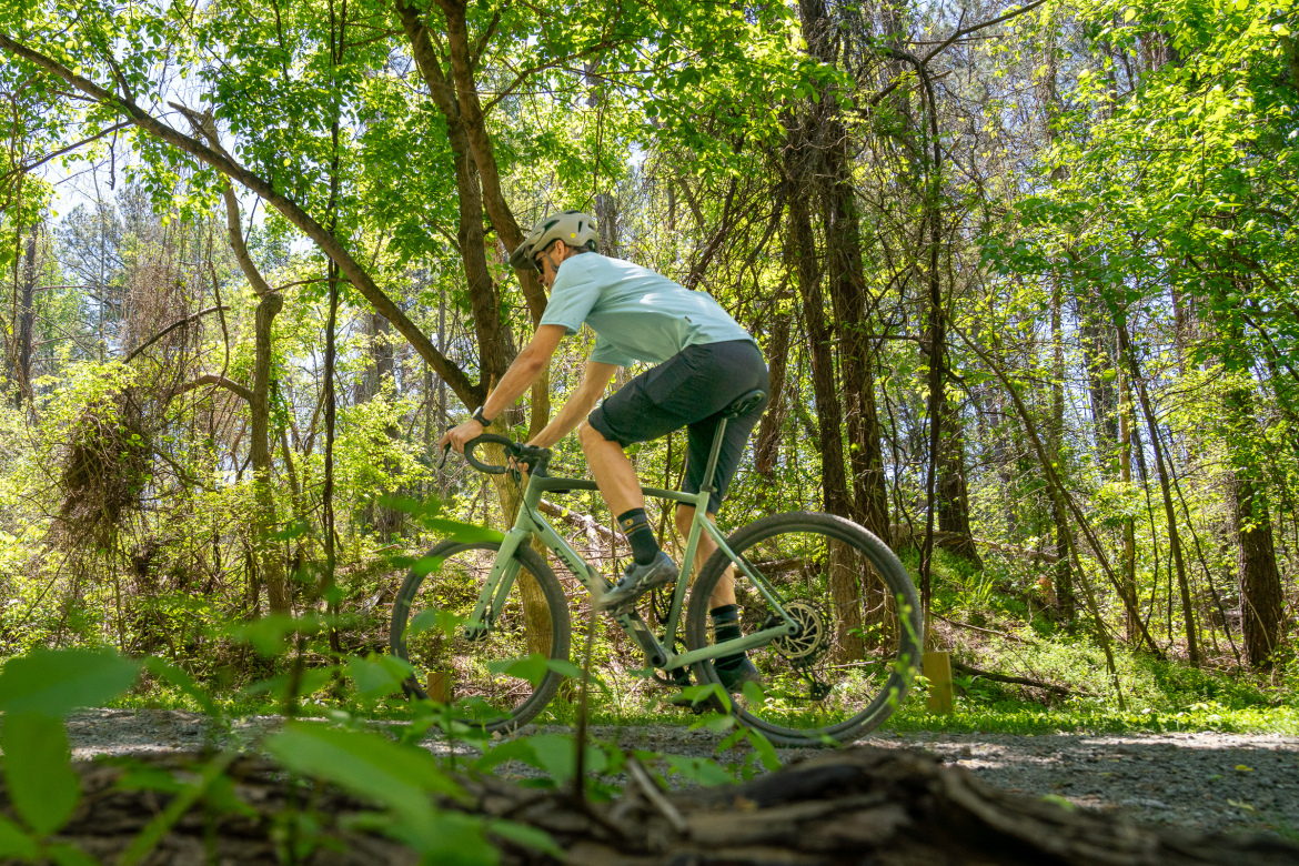 A cyclist riding a light green bike along a gravel path in a lush, green forest, surrounded by trees and natural foliage.