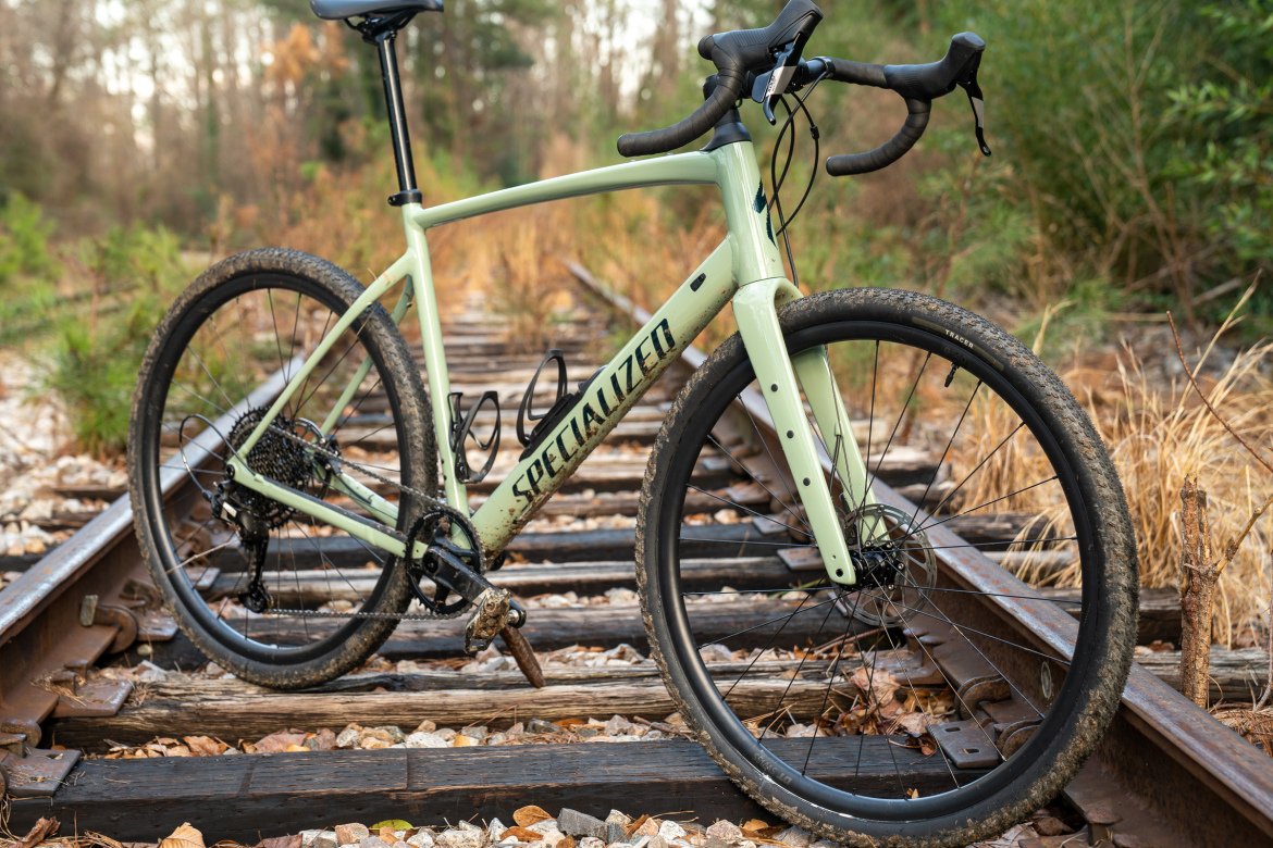 A light green gravel bike resting on weathered wooden train tracks, surrounded by overgrown grass and trees in the background. The bike features a sleek design with thin tires and drop handlebars, showcasing its readiness for outdoor adventures.