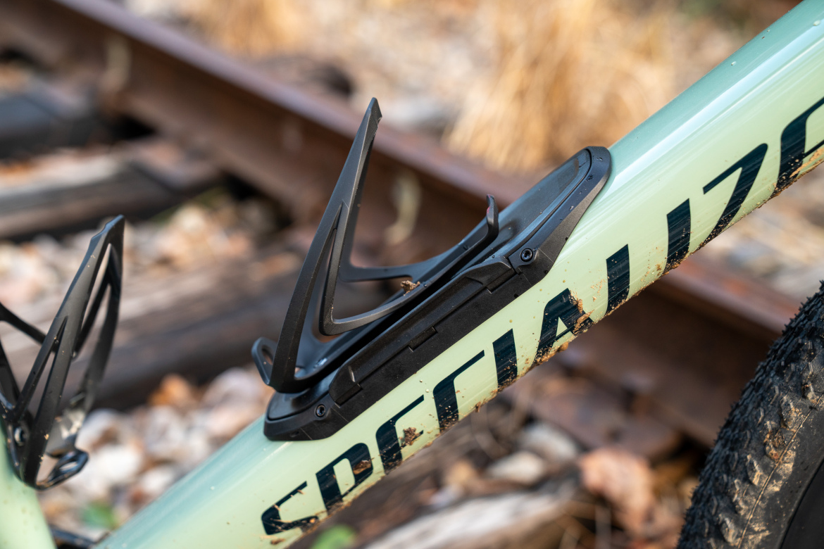 Close-up of a bicycle frame featuring a black water bottle cage mounted to the downtube, with the word "Specialized" visible on the frame. The background shows a rustic scene with wooden train tracks and autumn foliage.